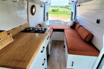 The interior of a neatly designed van conversion, showcasing a cozy, wood-accented kitchenette with a gas stove on the left and a narrow aisle leading to a seating area with orange cushions on the right. Large windows display a scenic view of a lush, green landscape outside.