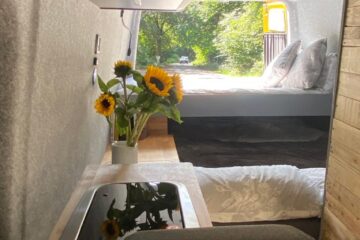 Interior of a campervan featuring a cozy bed with gray pillows at the back. A small wooden table with a vase holding sunflowers is next to the bed. The kitchen area includes a sleek black stovetop. Greenery is visible through the open rear doors, suggesting a park or natural setting outside.