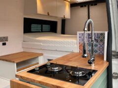 Interior of a modern camper van with wooden countertops. The foreground shows a compact kitchen area with a gas stove, sleek faucet, and colorful tiled backsplash. In the background, there is a neatly made bed with white bedding, and overhead cabinets for storage.