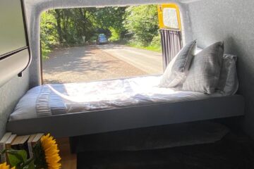 The image shows the interior of a parked camper van, viewed from the back. There's a neatly made bed with grey pillows and a blanket. Sunlight streams in, illuminating the bed. Outside the open back door, a tree-lined road is visible. A small sunflower bouquet and books are in the corner.