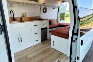 The image shows the interior of a white camper van converted into a cozy living space. It features white cabinets and drawers, a small sink with a black faucet, a wooden countertop, a small stove, a mini-fridge, and a cushioned seating area with a table. The back doors are open, revealing a scenic view outside.