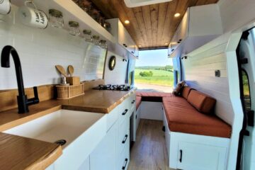 Interior of a converted van with a kitchen area featuring a white sink, black faucet, wooden countertops, and shelves holding jars and hanging mugs. A cozy seating area with orange cushions is at the back, with large windows framing a scenic view of a green countryside.