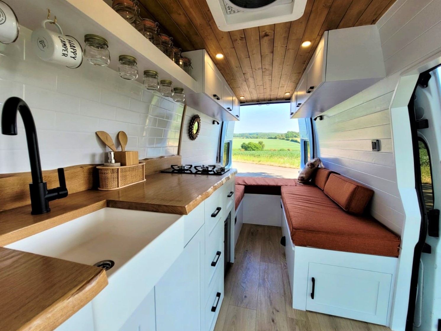 Interior of a converted van with a kitchen area featuring a white sink, black faucet, wooden countertops, and shelves holding jars and hanging mugs. A cozy seating area with orange cushions is at the back, with large windows framing a scenic view of a green countryside.