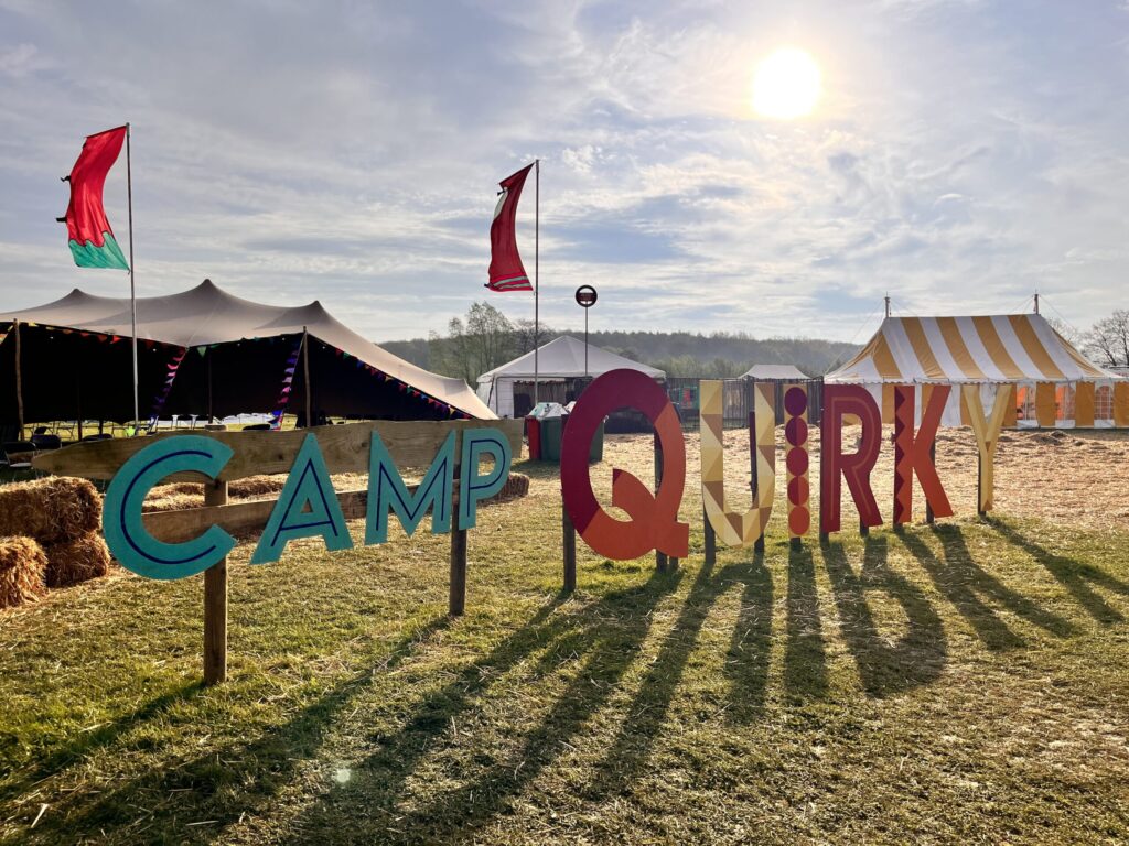 Sunlit outdoor festival scene with large colorful letters spelling "CAMP QUIRK" on the grass. Tents with bunting and flags are in the background, including a yellow-striped tent and a larger black tent. The sky is blue, slightly cloudy, and the sun is low, casting long shadows.