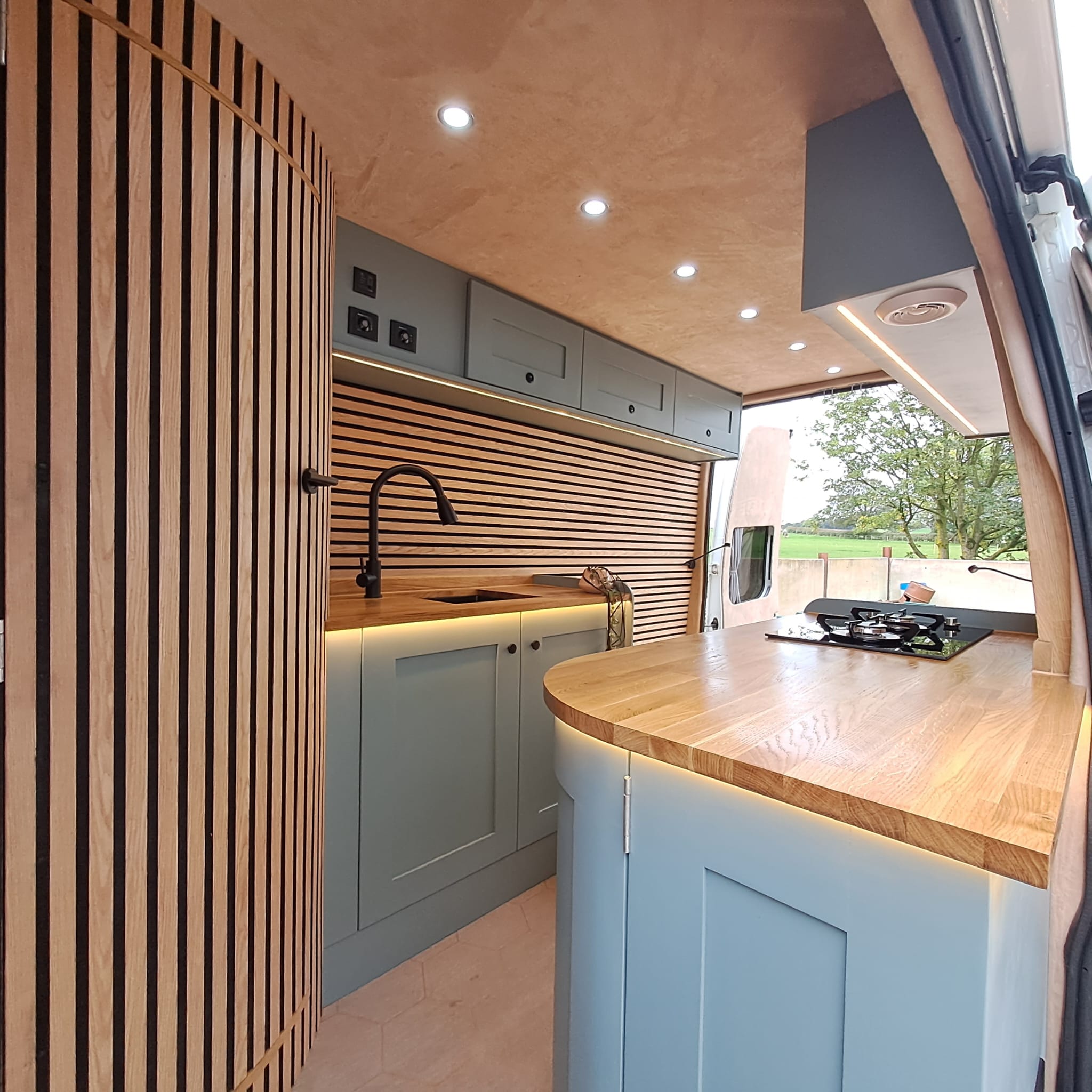 Interior of a stylish camper van with light wood paneling, soft lighting, and modern finishes. Features include a small kitchen area with a sink, stovetop, and wooden countertops. Overhead cabinets provide storage. A large window offers a view of trees outside.