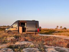 A grey van is parked on a rocky, grassy landscape under a clear sky during sunset. The van’s side door is open, revealing part of its interior. The serene environment includes patches of shrubs and distant trees, all bathed in warm, golden light from the setting sun.