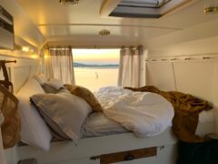 A cozy camper van interior with an unmade bed covered in white and beige linens, facing the back. The van doors are open, revealing a scenic view of a calm body of water and a sunset. Curtains frame the open doorway, and soft lighting creates a warm ambiance inside.