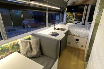 A well-lit, modern camper van interior features a small kitchen with a sink, faucet, and counter space on the left. Adjacent is a cushioned seating area with a table. The back has a cozy bed with polka dot bedding. Light wood flooring and large windows with greenery outside complete the space.