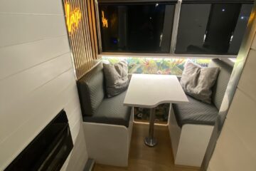 A cozy RV interior featuring a small dining area with two cushioned bench seats and a white table in between. The back wall has colorful, floral wallpaper and a neon sign. The wall on the left is white with a black heater, while the right side has large windows with black shades.