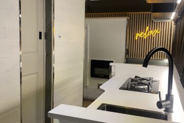 A modern RV interior features sleek white cabinetry and walls. At the center is a compact kitchen with a gas stove, sink, and black faucet. A neon sign reading "relax" is illuminated in the background. There's also a small electric fireplace under the sign and a bed with a white mattress in the foreground.