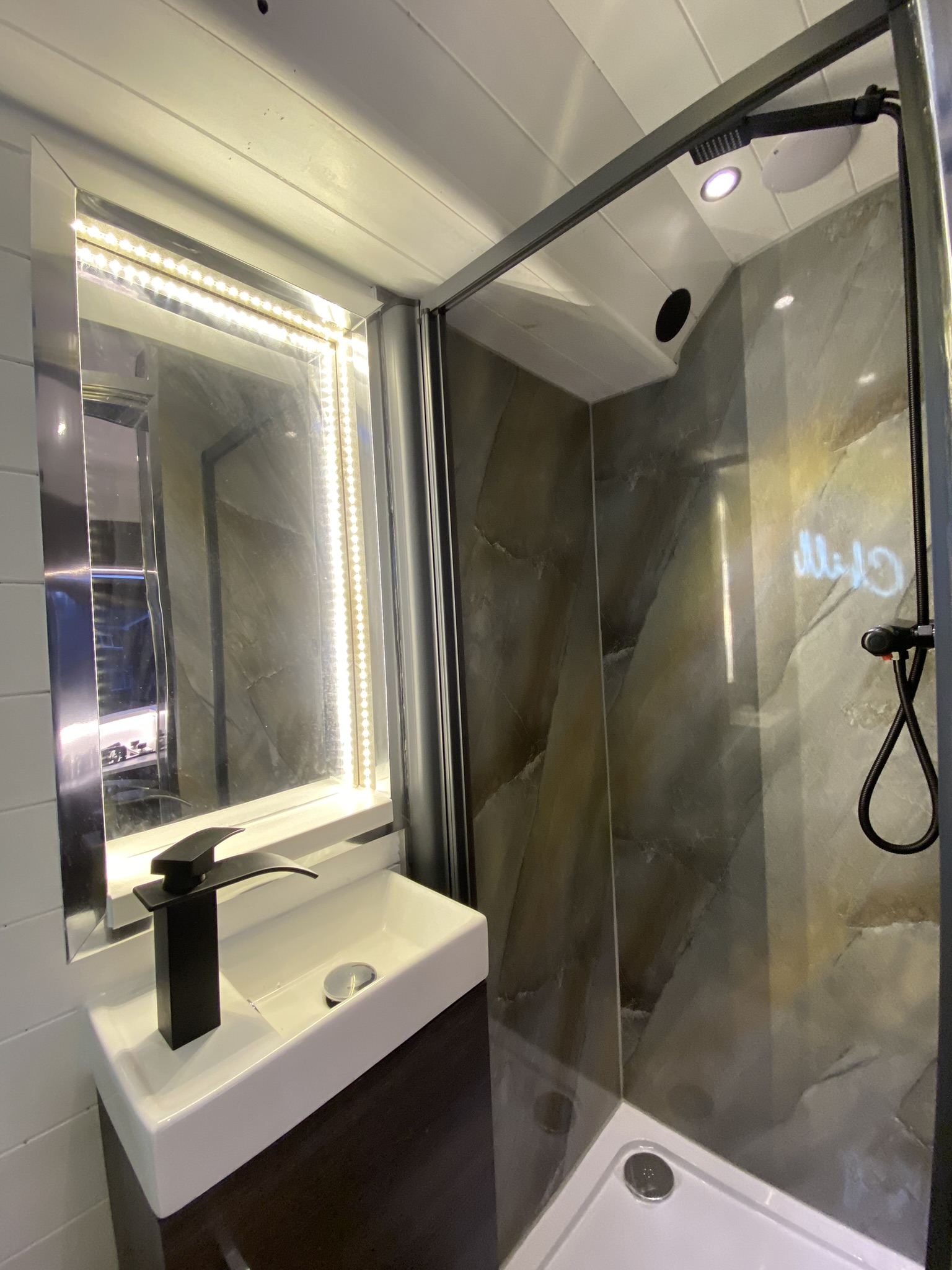 A modern bathroom features a sleek vanity with a rectangular sink and a black faucet. Above the sink is a large, illuminated mirror with LED lighting. Next to the vanity, there is a glass-enclosed shower with dark, textured tile walls and a black adjustable showerhead.