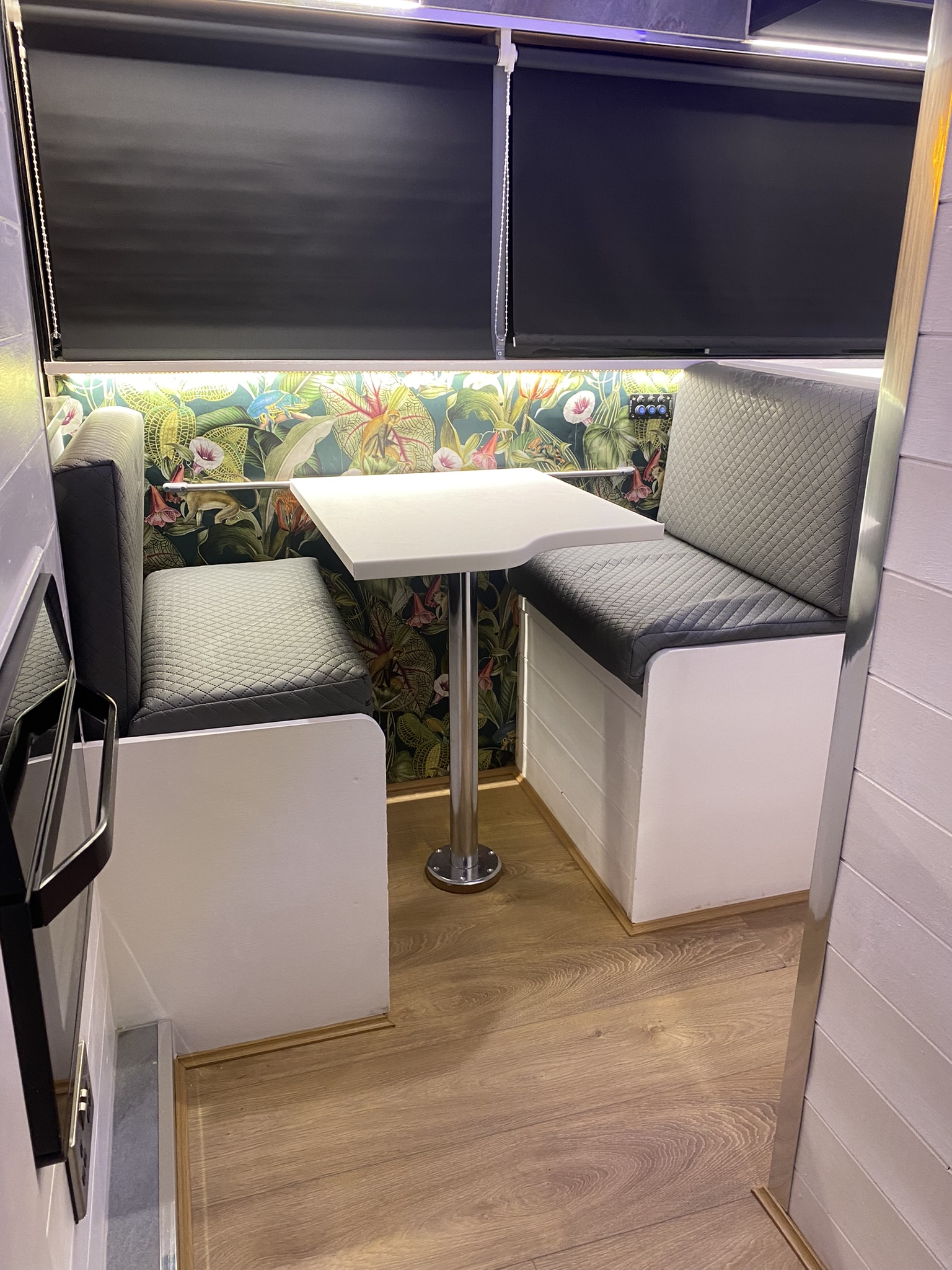 A cozy dining nook in an RV or tiny home, featuring a small square table with two quilted gray booth seats on either side. The walls are adorned with vibrant floral and tropical leaf wallpaper. The windows are covered with closed black blinds, and the floor is a light brown wood.