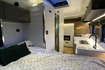 The image shows the interior of a modern, well-lit camper van. There is a bed with a white and green leaf-patterned duvet on the left, a small kitchen with a sink and stove on the right, and a vertical storage cabinet in the back. An open skylight allows natural light to enter.