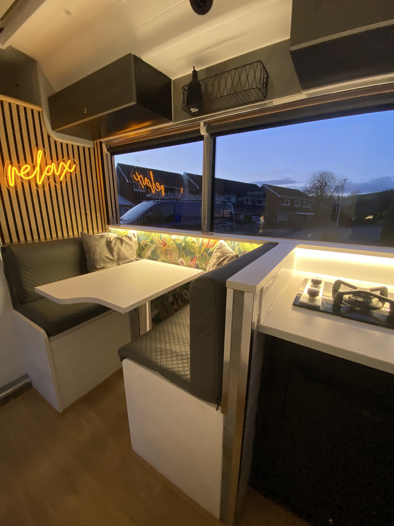 The interior of a modern camper van features a cozy dining area with a white table and two bench seats upholstered in grey quilted fabric. A neon 