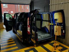 A blue van with its side and rear doors open is undergoing maintenance in a garage. The interior paneling has been removed, exposing the metallic framework. The van is parked on a floor with yellow and black striped markings. A tripod light stand is positioned near the rear of the van.