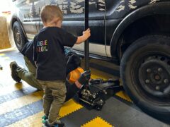 A young child stands next to a black vehicle holding a car jack handle, assisting an adult who is lying on their back underneath the car. The child wears a black shirt with colorful text, and the scene takes place on a black-and-yellow checkered floor in a garage.