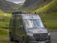 A gray Mercedes-Benz Sprinter van is parked on a grassy area near a winding road in a picturesque, mountainous landscape. The van has a roof rack equipped with storage and a ladder on its side. The sky is cloudy, casting a dramatic backdrop over the green hills and rugged terrain.