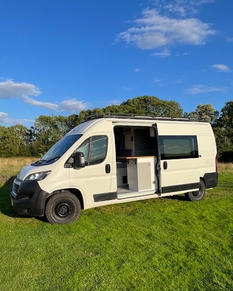 The main image portrays a white campervan with its side door open, showcasing a compact but organised interior. Parked on a green grassy field under a clear blue sky and equipped with black all-terrain tyres, this vehicle is nestled among trees, ideal for off-grid adventures. The weather is sunny with a few scattered clouds.