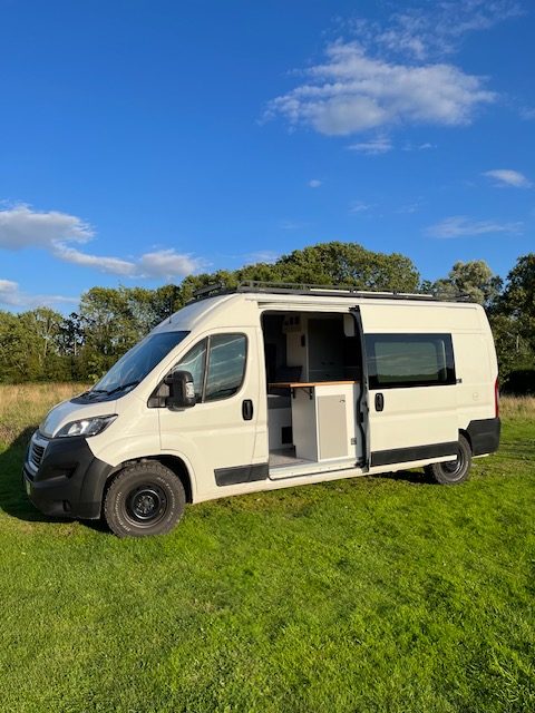 The main image portrays a white campervan with its side door open, showcasing a compact but organised interior. Parked on a green grassy field under a clear blue sky and equipped with black all-terrain tyres, this vehicle is nestled among trees, ideal for off-grid adventures. The weather is sunny with a few scattered clouds.