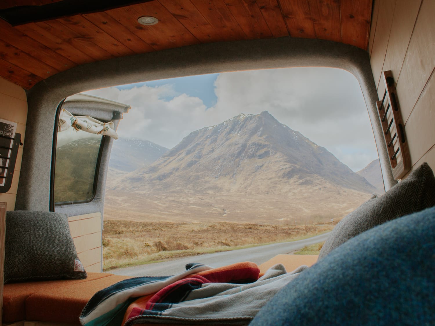 Inside a cosy campervan with a wooden interior, you can enjoy a scenic landscape featuring a large, rugged mountain under a cloudy blue sky. Blankets and cushions near the open door offer comfort and relaxation—the ideal setting for your campervan hire adventure away from Glasgow's bustling streets.